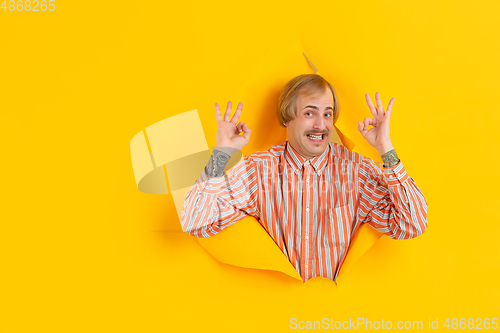 Image of Cheerful young man poses in torn yellow paper hole background, emotional and expressive