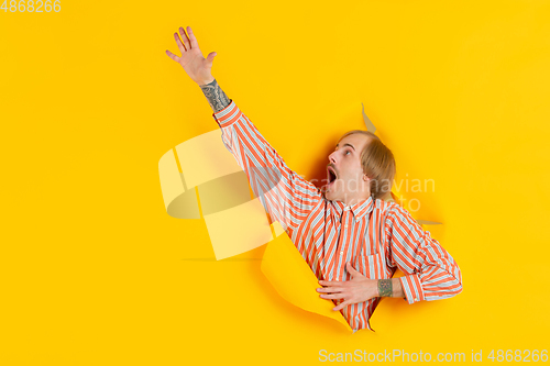 Image of Cheerful young man poses in torn yellow paper hole background, emotional and expressive