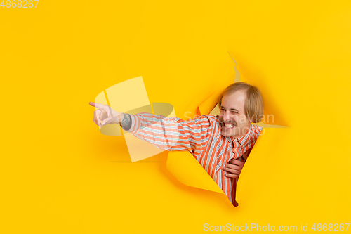 Image of Cheerful young man poses in torn yellow paper hole background, emotional and expressive