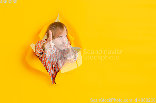 Image of Cheerful young man poses in torn yellow paper hole background, emotional and expressive