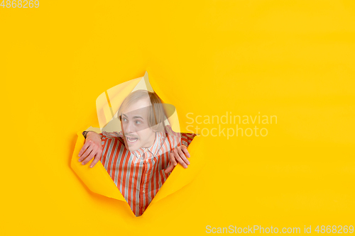 Image of Cheerful young man poses in torn yellow paper hole background, emotional and expressive