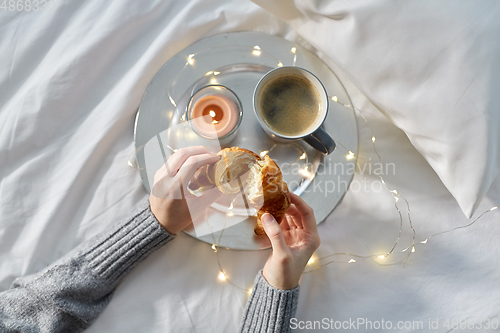 Image of hands of woman eating croissant with coffee in bed