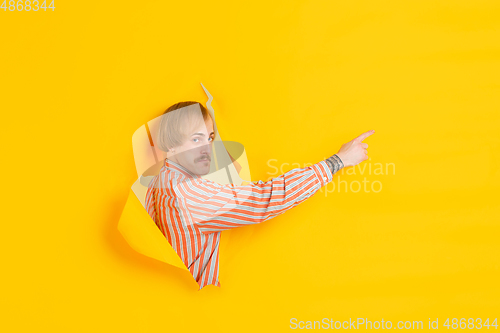 Image of Cheerful young man poses in torn yellow paper hole background, emotional and expressive