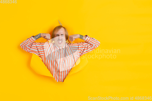 Image of Cheerful young man poses in torn yellow paper hole background, emotional and expressive