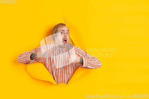 Image of Cheerful young man poses in torn yellow paper hole background, emotional and expressive