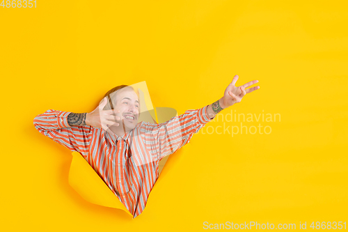 Image of Cheerful young man poses in torn yellow paper hole background, emotional and expressive