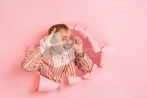 Image of Cheerful young man poses in torn coral paper hole background, emotional and expressive