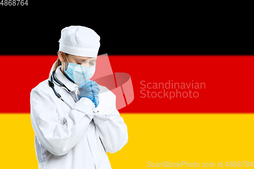 Image of Female young doctor with stethoscope and face mask praying for God with Germany national flag on background