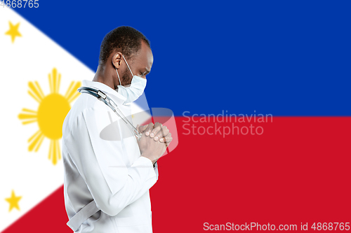 Image of Young doctor with stethoscope and face mask praying for God with Philippines national flag on background