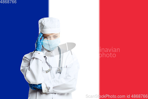 Image of Female young doctor with stethoscope and face mask praying for God with France national flag on background