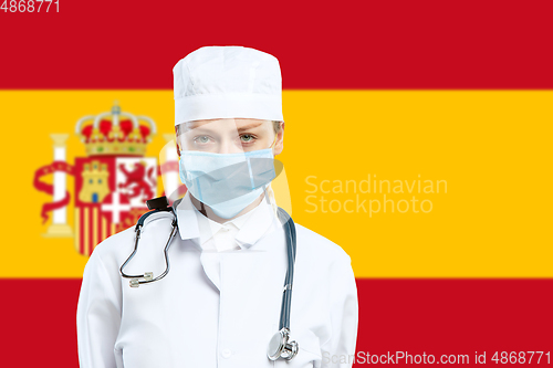 Image of Female young doctor with stethoscope and face mask praying for God with Spain national flag on background