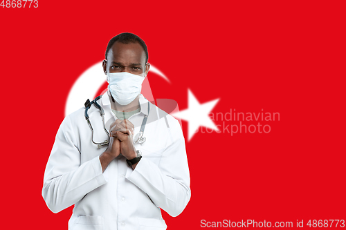 Image of Young doctor with stethoscope and face mask praying for God with Turkey national flag on background