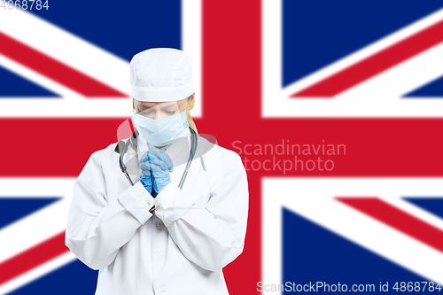 Image of Female young doctor with stethoscope and face mask praying for God with United Kingdom national flag on background