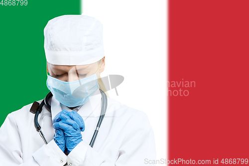 Image of Female young doctor with stethoscope and face mask praying for God with Italy national flag on background