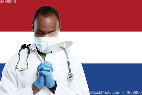 Image of Young doctor with stethoscope and face mask praying for God with Netherlands national flag on background