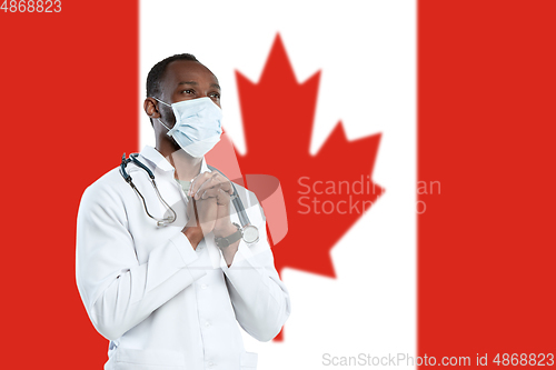 Image of Young doctor with stethoscope and face mask praying for God with Canada national flag on background