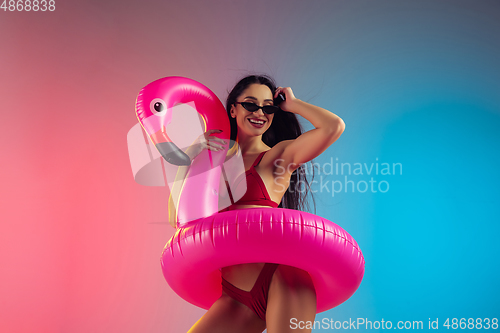 Image of Fashion portrait of young fit and sportive woman with rubber flamingo in stylish red swimwear on gradient background. Perfect body ready for summertime.