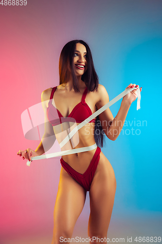 Image of Close up of young fit and sportive woman with measurer in stylish red swimwear on gradient background. Perfect body ready for summertime.