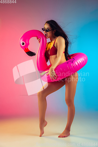 Image of Fashion portrait of young fit and sportive woman with rubber flamingo in stylish red swimwear on gradient background. Perfect body ready for summertime.