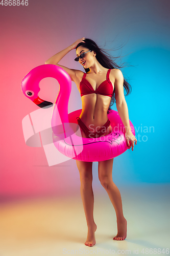 Image of Fashion portrait of young fit and sportive woman with rubber flamingo in stylish red swimwear on gradient background. Perfect body ready for summertime.