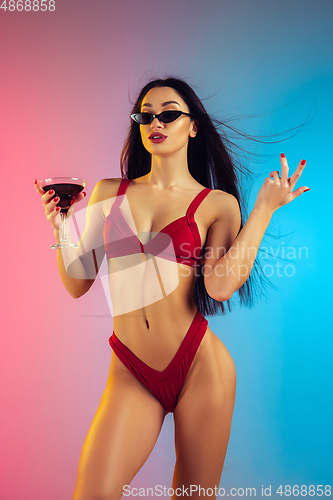 Image of Fashion portrait of young fit and sportive woman with cocktail in stylish red luxury swimwear on gradient background. Perfect body ready for summertime.