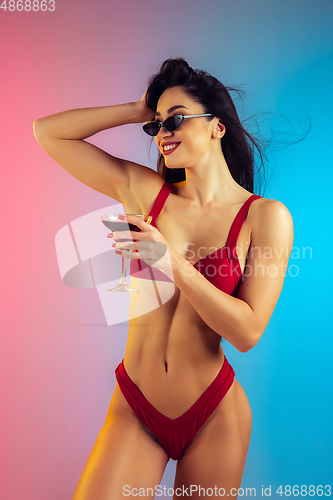 Image of Fashion portrait of young fit and sportive woman with cocktail in stylish red luxury swimwear on gradient background. Perfect body ready for summertime.