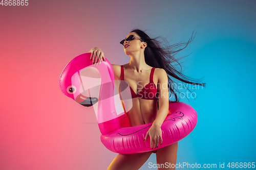 Image of Fashion portrait of young fit and sportive woman with rubber flamingo in stylish red swimwear on gradient background. Perfect body ready for summertime.