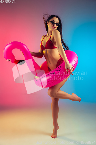 Image of Fashion portrait of young fit and sportive woman with rubber flamingo in stylish red swimwear on gradient background. Perfect body ready for summertime.