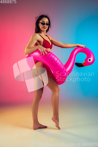 Image of Fashion portrait of young fit and sportive woman with rubber flamingo in stylish red swimwear on gradient background. Perfect body ready for summertime.