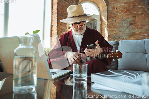 Image of Mature senior older man during quarantine, realizing how important stay at home during virus outbreak, giving concert of taking online lessons of guitar playing