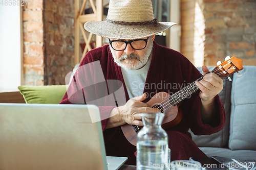 Image of Mature senior older man during quarantine, realizing how important stay at home during virus outbreak, giving concert of taking online lessons of guitar playing