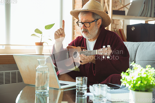 Image of Mature senior older man during quarantine, realizing how important stay at home during virus outbreak, giving concert of taking online lessons of guitar playing