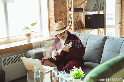 Image of Mature senior older man during quarantine, realizing how important stay at home during virus outbreak, giving concert of taking online lessons of guitar playing