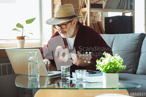 Image of Mature senior older man during quarantine, realizing how important stay at home during virus outbreak, giving concert of taking online lessons of guitar playing