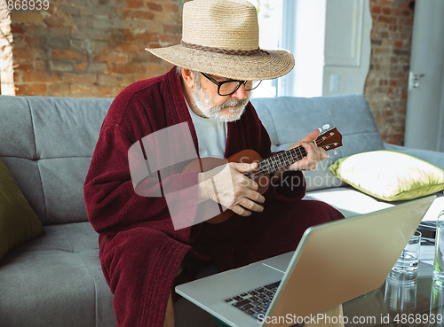 Image of Mature senior older man during quarantine, realizing how important stay at home during virus outbreak, giving concert of taking online lessons of guitar playing