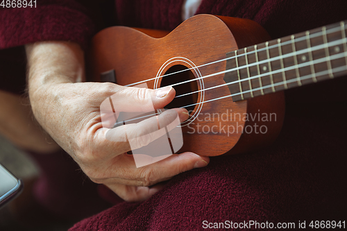 Image of Mature senior older man during quarantine, virus outbreak, giving concert of taking online lessons of guitar playing, close up of hands