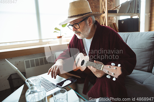 Image of Mature senior older man during quarantine, realizing how important stay at home during virus outbreak, giving concert of taking online lessons of guitar playing