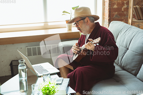 Image of Mature senior older man during quarantine, realizing how important stay at home during virus outbreak, giving concert of taking online lessons of guitar playing