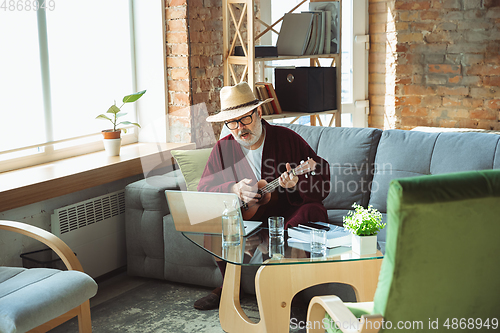 Image of Mature senior older man during quarantine, realizing how important stay at home during virus outbreak, giving concert of taking online lessons of guitar playing