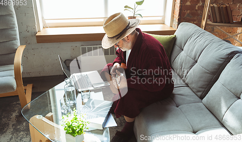 Image of Mature senior older man during quarantine, realizing how important stay at home during virus outbreak, giving concert of taking online lessons of guitar playing