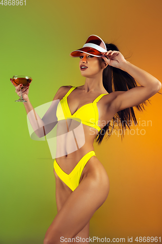 Image of Fashion portrait of young fit and sportive woman with cocktail in stylish yellow luxury swimwear on gradient background. Perfect body ready for summertime.