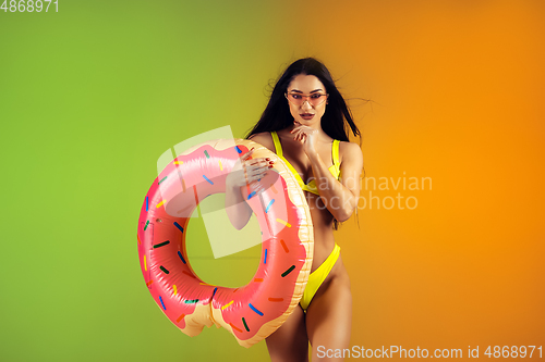 Image of Fashion portrait of young fit and sportive woman with rubber donut in stylish yellow swimwear on gradient background. Perfect body ready for summertime.