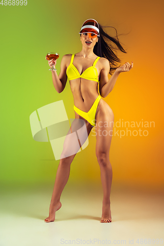 Image of Fashion portrait of young fit and sportive woman with cocktail in stylish yellow luxury swimwear on gradient background. Perfect body ready for summertime.