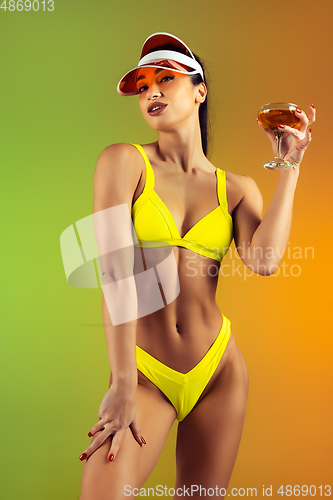 Image of Fashion portrait of young fit and sportive woman with cocktail in stylish yellow luxury swimwear on gradient background. Perfect body ready for summertime.