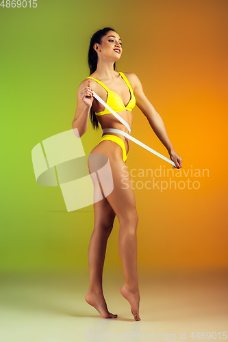 Image of Close up of young fit and sportive woman with measurer in stylish yellow swimwear on gradient background. Perfect body ready for summertime.