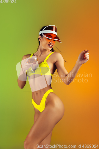 Image of Fashion portrait of young fit and sportive woman with cocktail in stylish yellow luxury swimwear on gradient background. Perfect body ready for summertime.