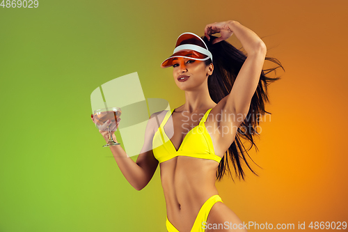 Image of Fashion portrait of young fit and sportive woman with cocktail in stylish yellow luxury swimwear on gradient background. Perfect body ready for summertime.