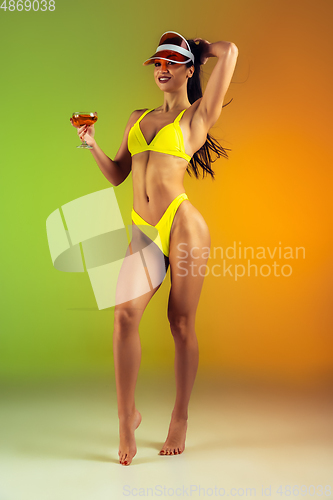 Image of Fashion portrait of young fit and sportive woman with cocktail in stylish yellow luxury swimwear on gradient background. Perfect body ready for summertime.