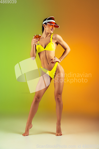 Image of Fashion portrait of young fit and sportive woman with cocktail in stylish yellow luxury swimwear on gradient background. Perfect body ready for summertime.