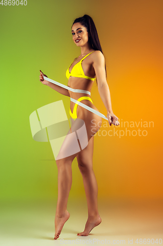 Image of Close up of young fit and sportive woman with measurer in stylish yellow swimwear on gradient background. Perfect body ready for summertime.